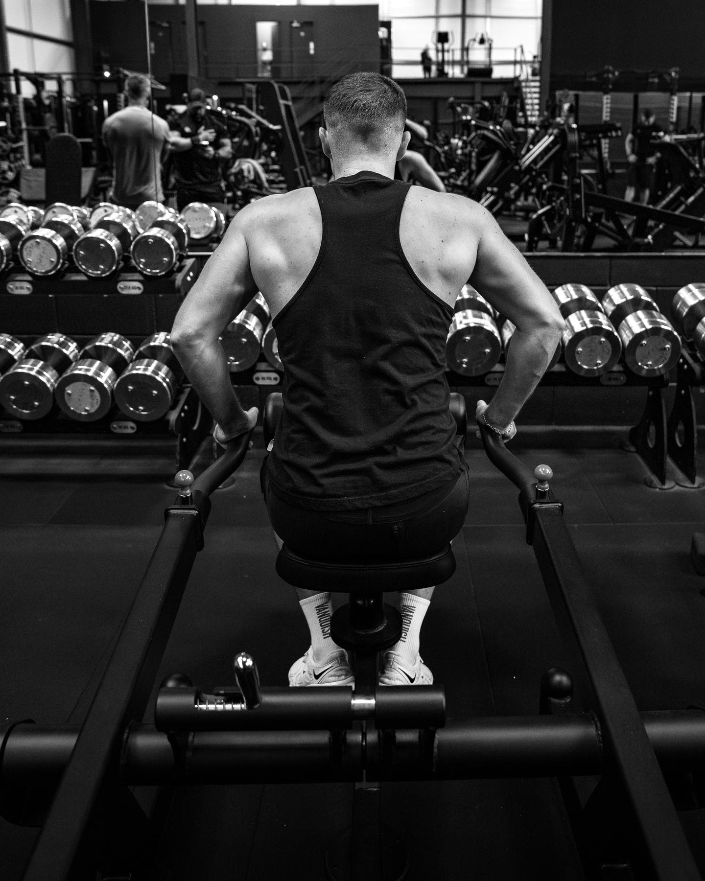A man, seen from behind, trains seated at a machine in front of a dumbbell rack.